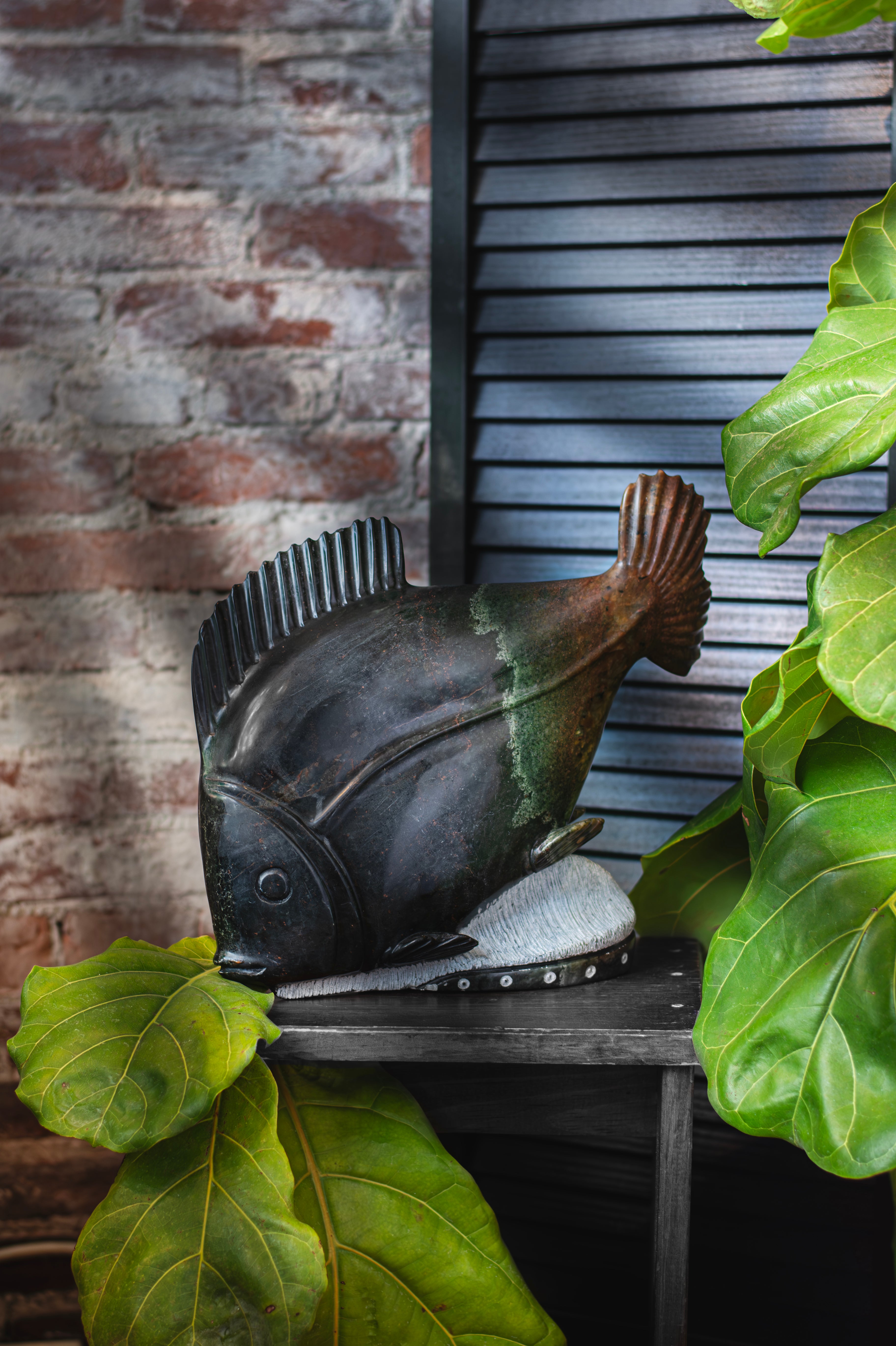 Image of fish sculpture on a black wooden bench in front of a brick wall and black shutter